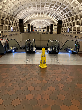 A picture of a set of escalators in a metro station. In front of the escalators there is one yellow wet floor cone. The escalators have a half wall running around the three other sides. In the background there are six wayfinding structures.